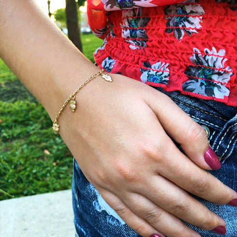 A person adorned in a floral top and the Owl Charms Bracelet and Anklet, an embodiment of wisdom and intuition in 14k gold-filled material, rests their hand on their denim-clad leg, highlighting dark pink nail polish.